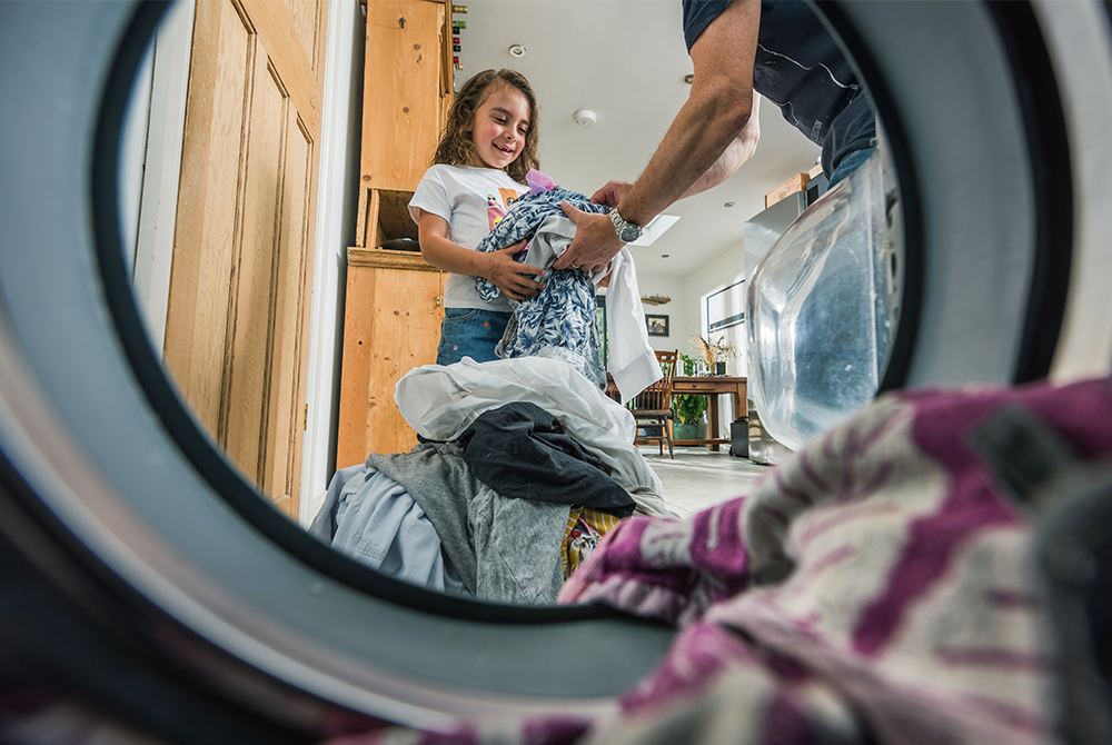 Small child loading clothes into a washing machine with help from an adult
